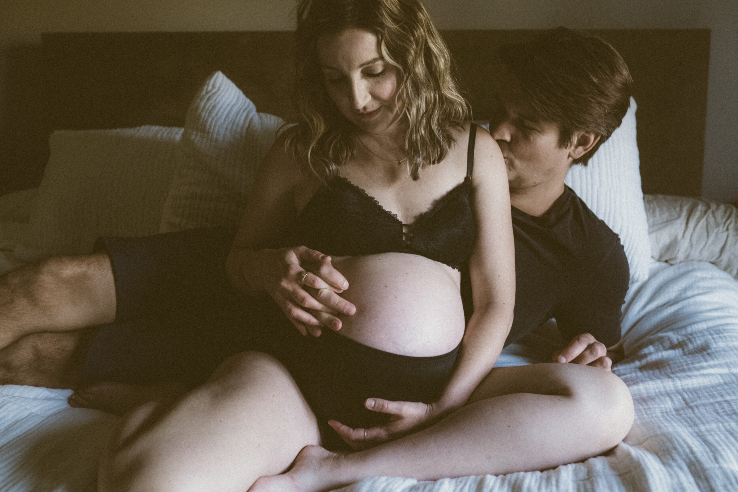 Pregnant woman and her husband sitting together on a bed, sharing a quiet, intimate moment during an in-home maternity session in Kansas City.